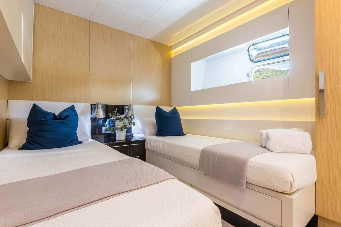 Modern bedroom aboard Arcadia 85 yacht for charter, with twin beds, blue pillows, warm lights, and a flowered nightstand.