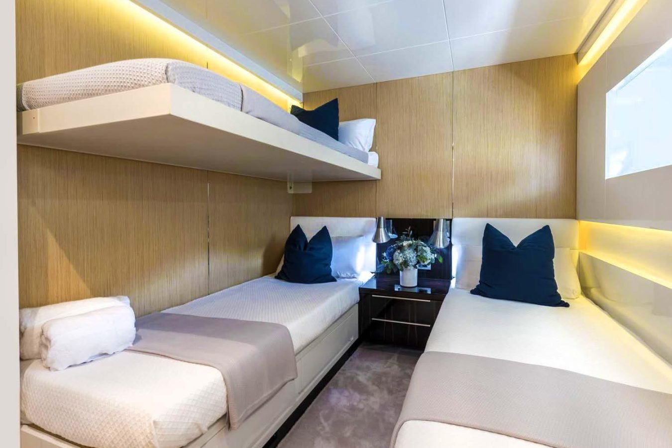 Modern bedroom on Dhamma II yacht for charter with twin beds, overhead bunk, blue pillows, and a flowered nightstand.