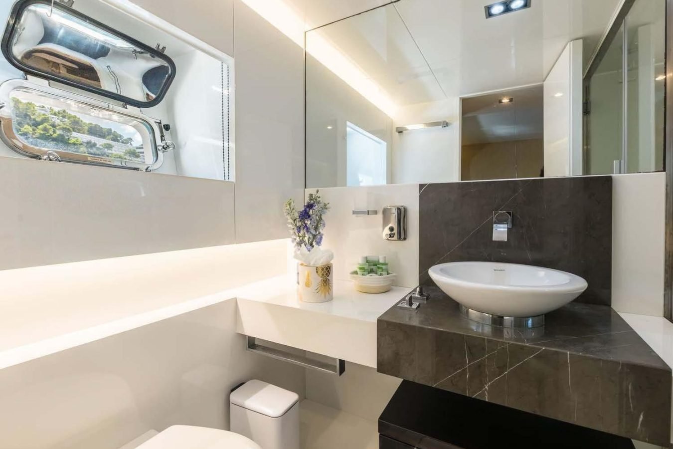 Modern bathroom aboard Arcadia 85 Dhamma II with round sink, large mirror, and vase on a sleek white counter.