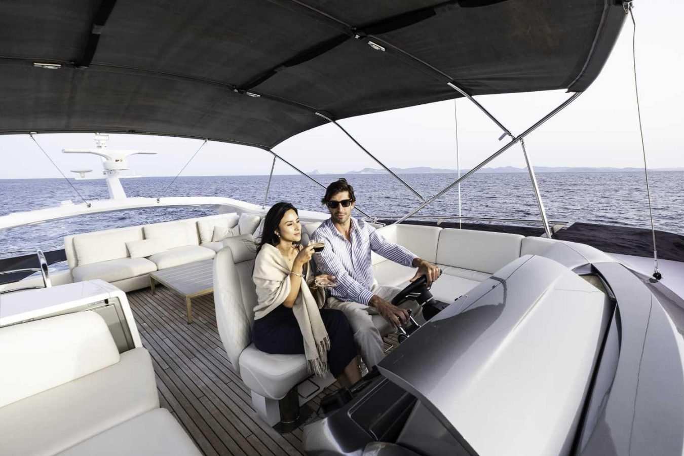A man steers the Princess S78 as a woman smiles beside him, holding a glass, with the sea glistening beyond.