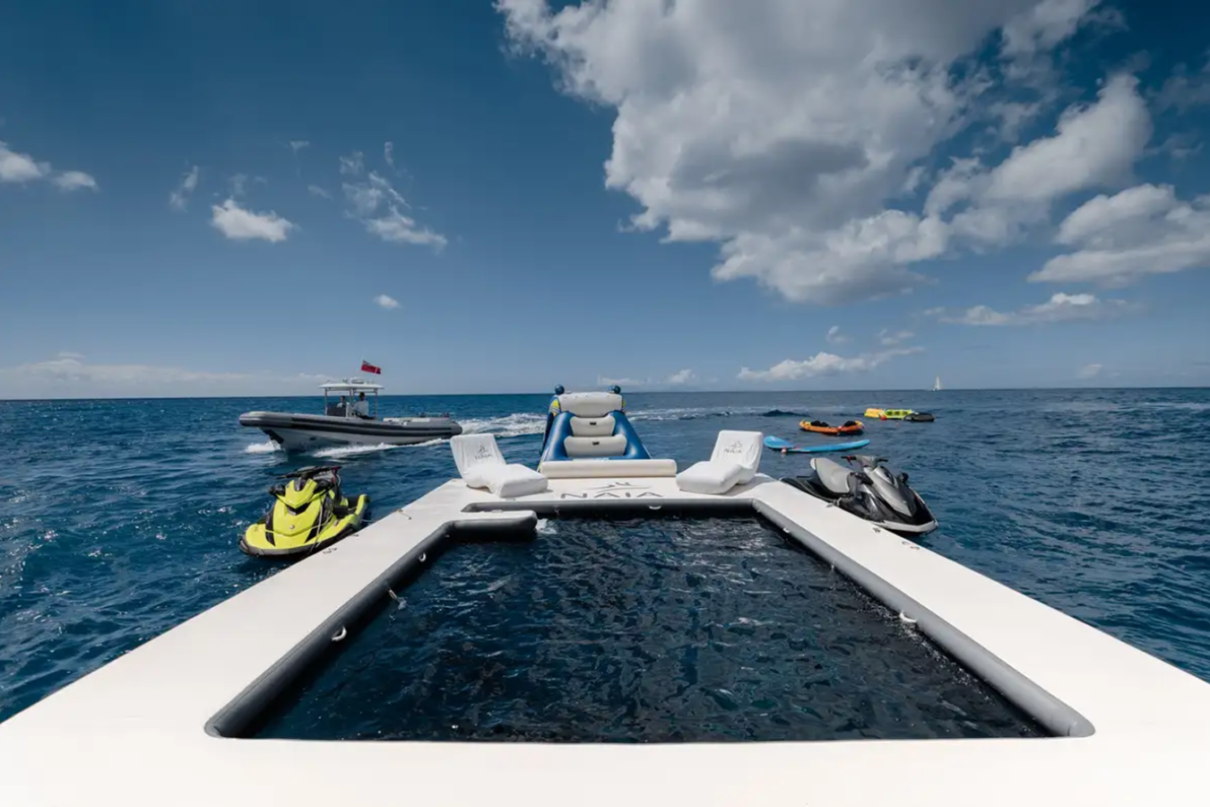 View from the back of Freire Naia Yacht for Charter with a floating pool, jet skis, and another boat on the blue ocean.