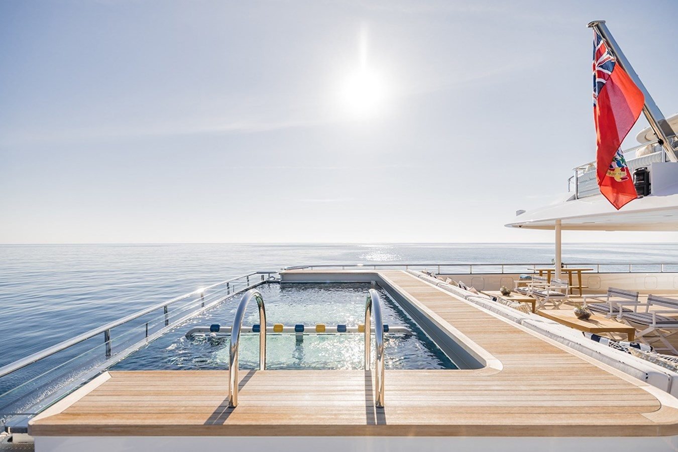 An Abeking & Rasmussen HBC Yacht for Charter deck features a pool, lounge chairs, and a flag under the bright sun.
