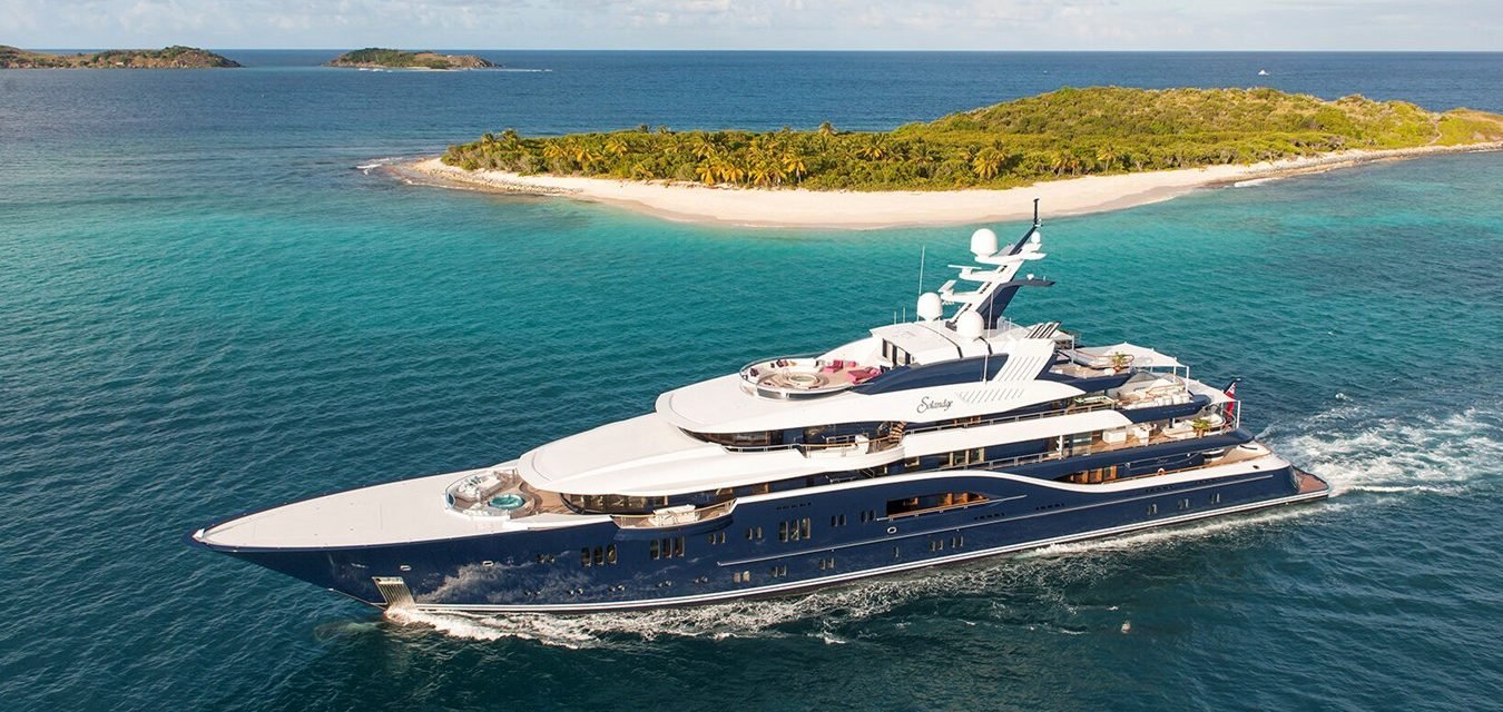 The Lurssen Solandge Yacht sails near a small tropical island with lush greenery and white sandy beaches.