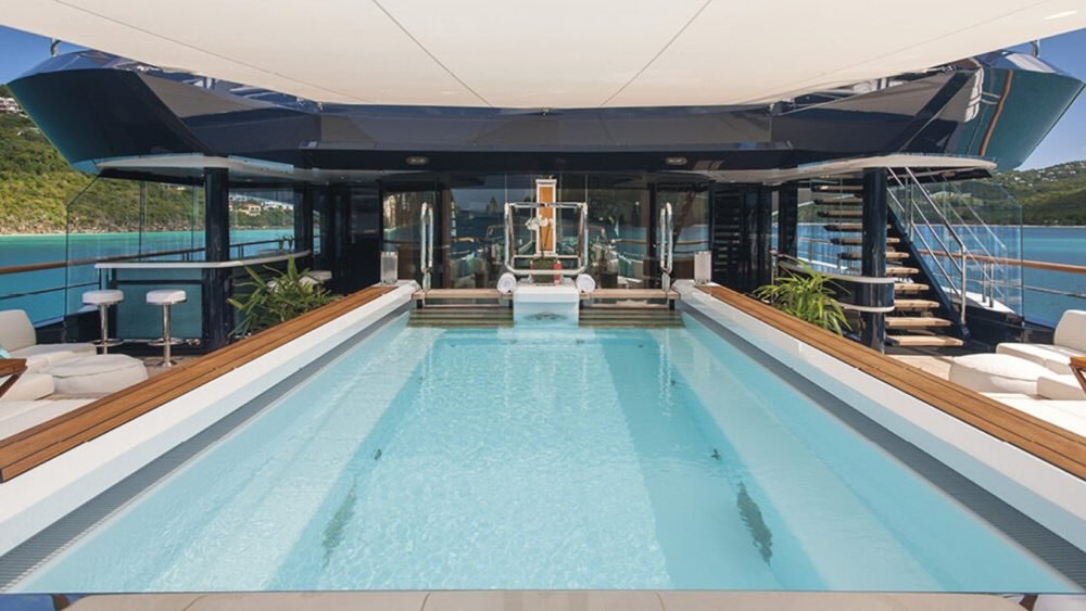 Luxury Lurssen Solandge Yacht deck with a central pool, lounge seating, bar stools, stairs, and ocean view.