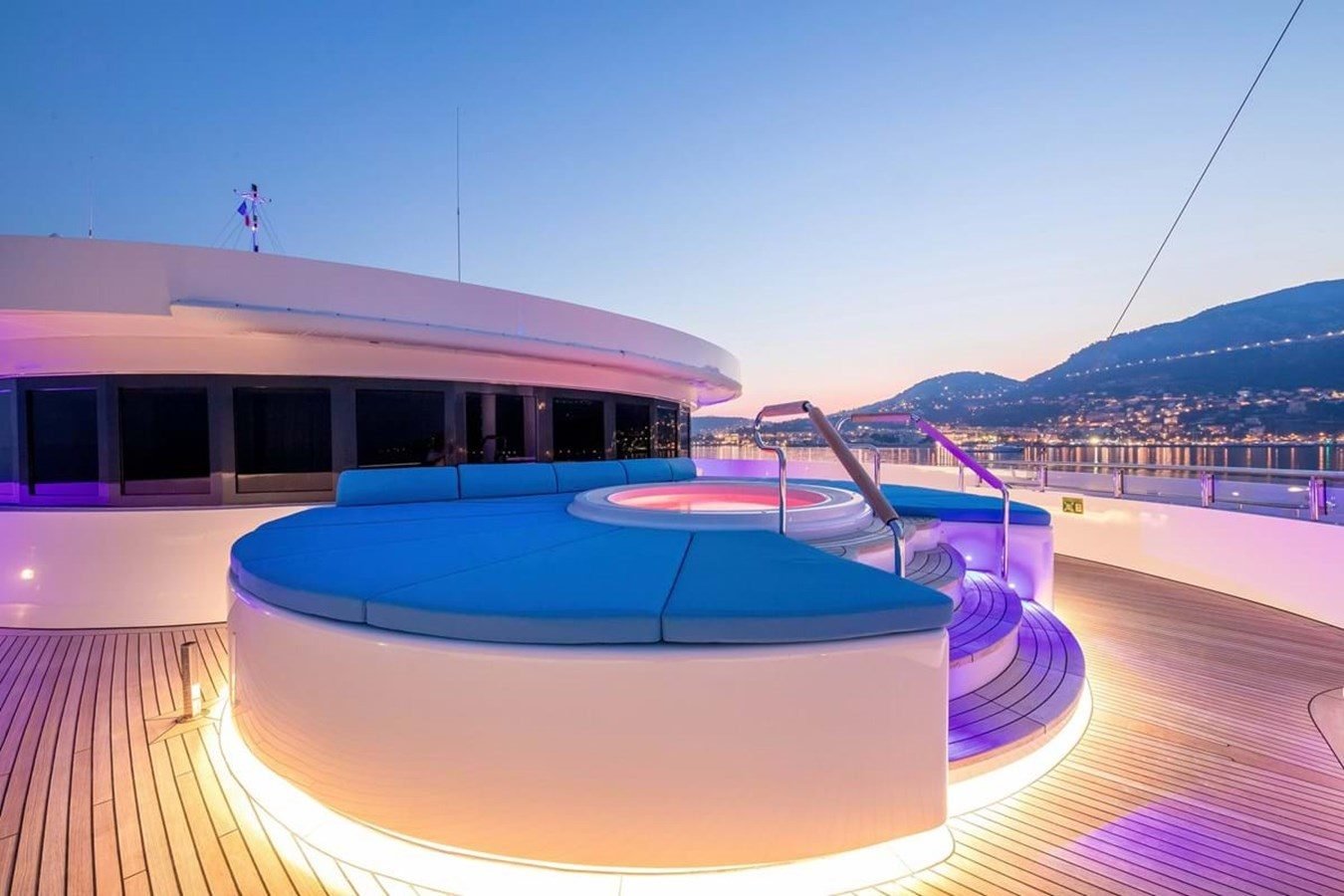 A luxury Neorion Moonlight II Yacht for Charter deck at sunset with a hot tub, blue cushions, and city lights on calm water.