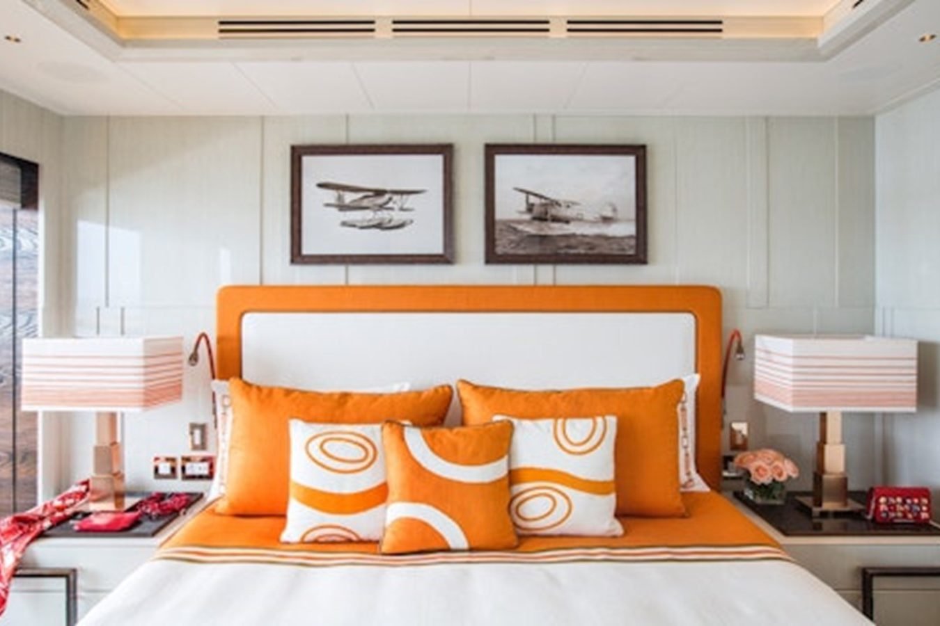 Modern bedroom with orange and white bed, patterned pillows, and vintage airplane photos evokes Lurssen CC-Summer Yacht style.