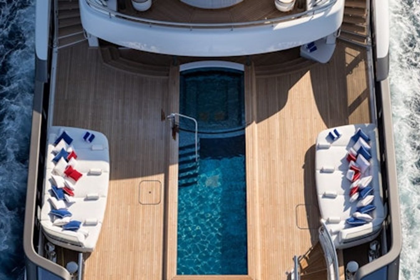 Aerial view of Lurssen CC-Summer Yacht for Charter, featuring a central pool and vibrant lounge areas with red, white, and blue pillows.