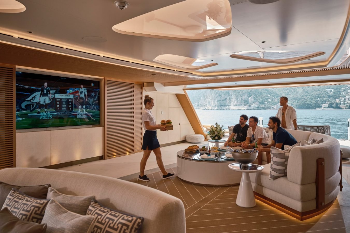 Four people watch football on a large TV aboard the Freire Renaissance Yacht for Charter as a server brings food; ocean views outside.
